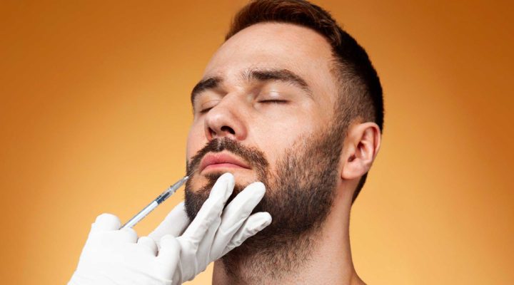 Close-up of a man with eyes closed as a gloved hand administers a syringe to his chin for a cosmetic facial injection on an orange background.”