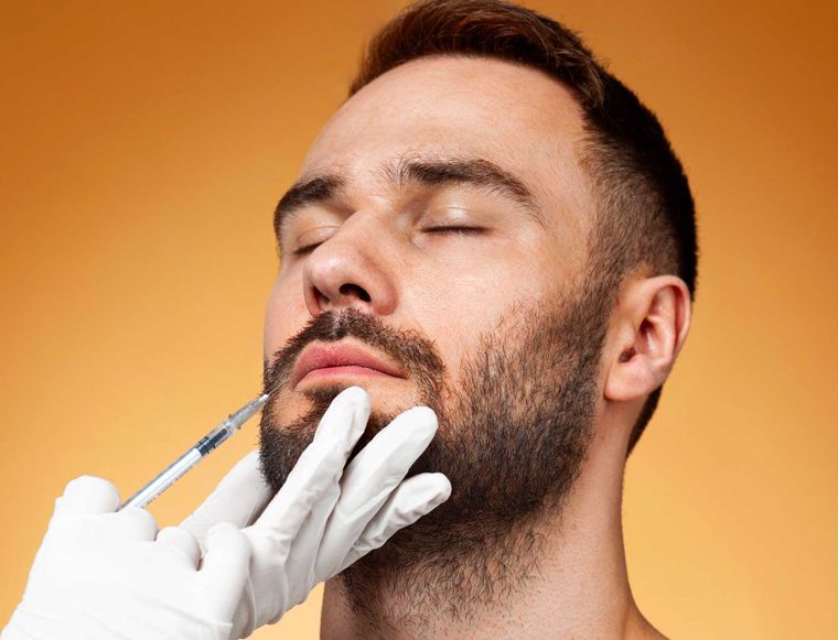 Close-up of a man with eyes closed as a gloved hand administers a syringe to his chin for a cosmetic facial injection on an orange background.”