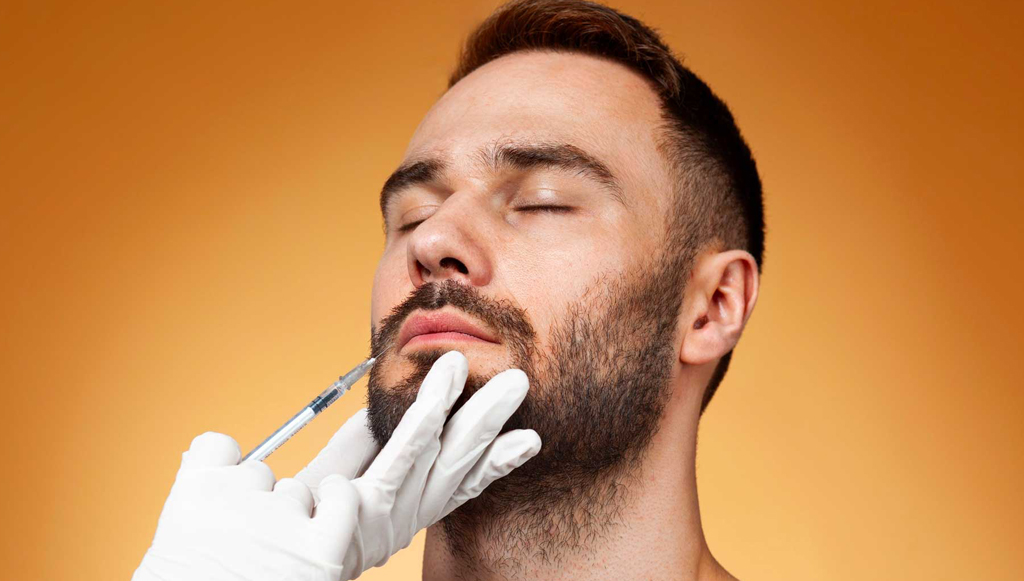 Close-up of a man with eyes closed as a gloved hand administers a syringe to his chin for a cosmetic facial injection on an orange background.”