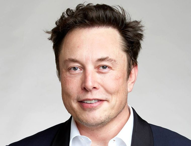 Portrait of a man in a dark blazer and white shirt smiling against a light gray background near-crop headshot
