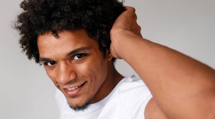 Portrait of a smiling man with curly hair wearing a white T-shirt, hand resting on the back of his head.