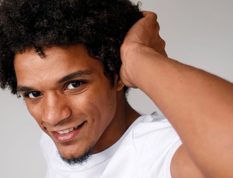 Smiling young man with curly hair and a white shirt, adjusting his hair with one hand against a neutral background.