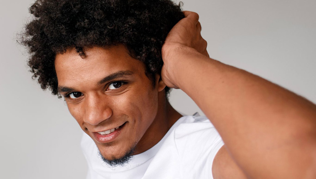 Smiling young man with curly hair and a white shirt, adjusting his hair with one hand against a neutral background.